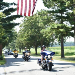 Line of motorcyclists riding under American flag on tree-lined street.