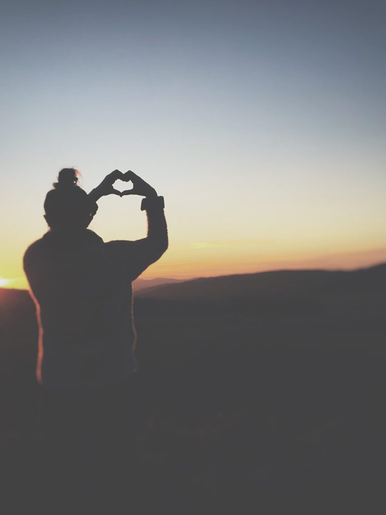 Silhouette of girl on mountain top holding her hands in a heart shape.